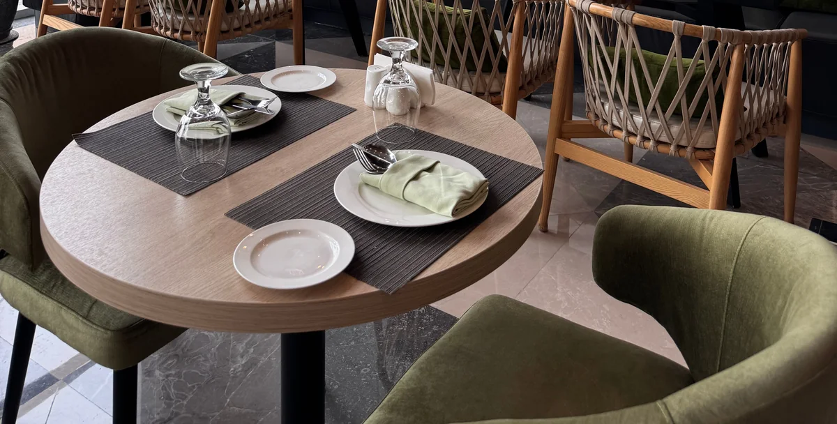 A round restaurant table set with plates and glassware, surrounded by green and woven chairs